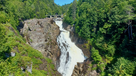 Beautiful waterfall powerfully cascades down a rocky gorge surrounded by lush green forest under a clear blue sky.の写真素材