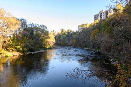 Humber River flowing through Etienne Brule Park, flanked by changing autumn foliage and distant buildings on the hillside, creating a serene urban nature landscape during fall seasonの写真素材