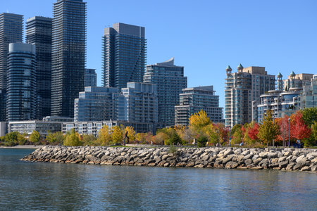 Etobicoke waterfront showing the modern skyline of residential high-rise buildings and colorful autumn trees lining the rocky shoreline under a clear blue sky, illustrating urban living by the lakeの写真素材