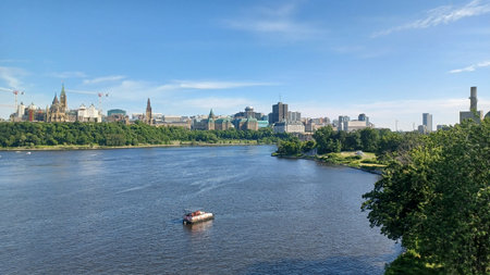 Ottawa, ON, Canada - August 31, 2025 the city skyline, featuring the Parliament buildings, stretches across the horizon with a wide river and a single boat in the foreground under a clear blue sky.のeditorial素材