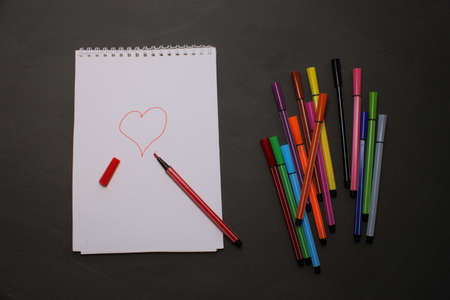 Markers of different colors with a white notebook on a black background. View from above. Colorful...