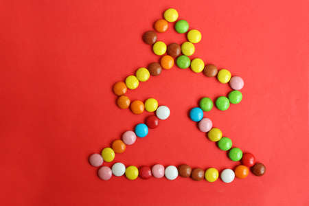 Chocolate small candies are laid out in the form of a Christmas tree on a red background. A symbol of the new year and Christmas. Top view. The candy is multi-colored. Copy space for text.の写真素材