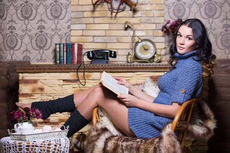 Brunette in blue sweater posing on the chair near a fireplace with bookの写真素材