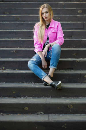 Beautiful blonde posing outdoors sitting on the stairs dressed in a pink jacket and pantsの写真素材