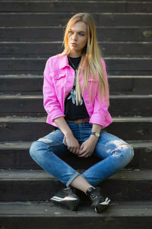 Nice blonde posing outdoors sitting on the stairs dressed in a pink jacket and pantsの写真素材