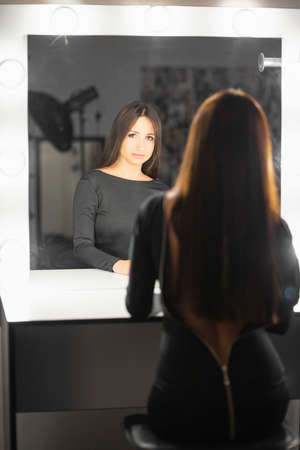 Portrait of a alluring lady posing in the studio sitting on a chair and looking into a mirror dressed in a dressの写真素材