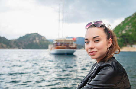 Portrait of a beautiful young brunette posing on a background of mountains, sea and a ship, wearing...