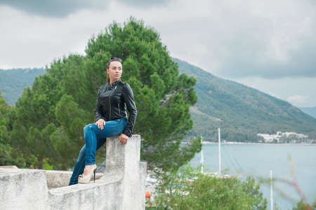 Young beautiful brunette on a background of mountains and the sea posing dressed in a jacket and...