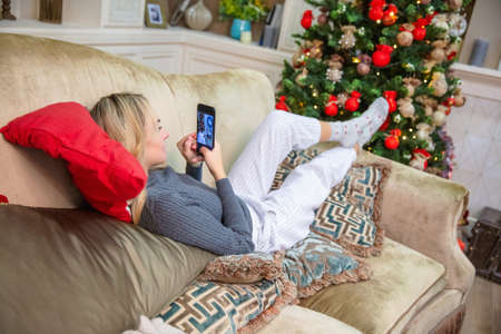 Beautiful young girl lies on a sofa and takes a selfie, dressed in pants and a sweater