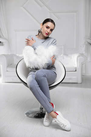 Beautiful brunette wearing a sweater with fur and jeans sitting on an armchairの写真素材