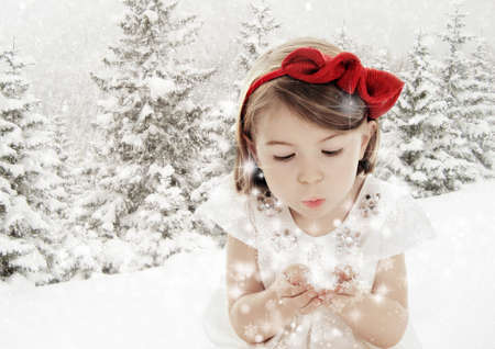 Beautiful little girl blowing snowflakes in white winter forest covered with snowの写真素材