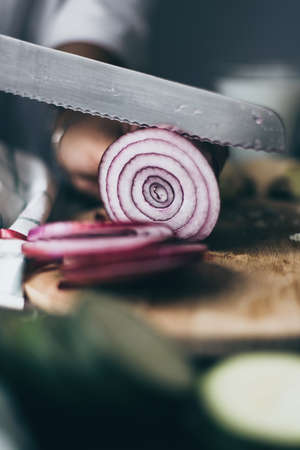 Onion being sliced with a knife. Artistic view of food. Tasty recipe. Delicious meal cooking. Warm colors and wooden cutting board. Brown background. Green cabbage on the front.の写真素材