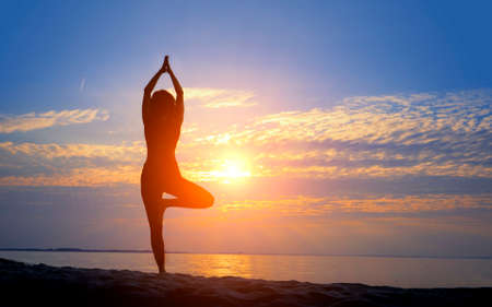 Female silhouette doing yoga asana at sunrise with hands raised to sunの写真素材