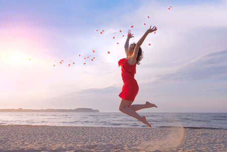 Jumping beautiful woman in red dress throwing rose petals on beach.の写真素材