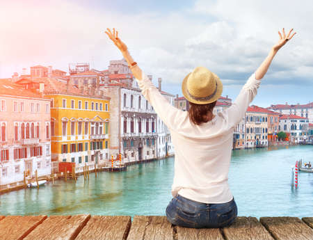 Woman traveler in hat with arms raised on bridge in Venice. Back view with copy spaceの写真素材