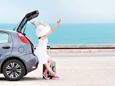 Woman traveler with raised arms sitting on hatchback car on beachの写真素材