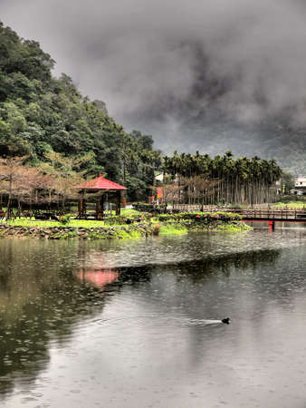 A lake with mountain, a pavilion, trees, and a duck on the surface の写真素材