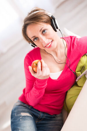 pretty young women on a sofa, listening music on her tablet computer and eating an appleの写真素材