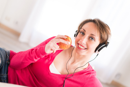 pretty young women on a sofa, listening music on her tablet computer and eating an appleの写真素材