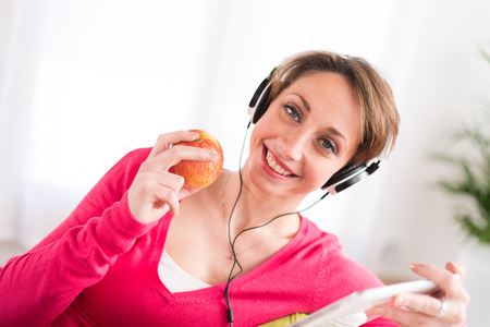 pretty young women on a sofa, listening music on her tablet computer and eating an appleの写真素材
