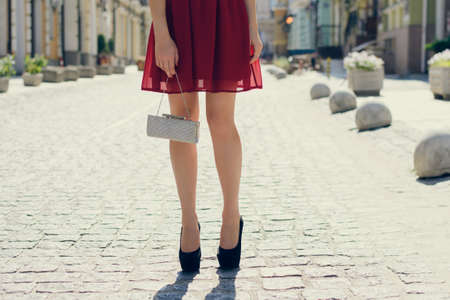 Beautiful young girl in red evening dress with bag in hands waiting for somebody in the street. Close up photo of woman's long legs in black  high-heelsの写真素材