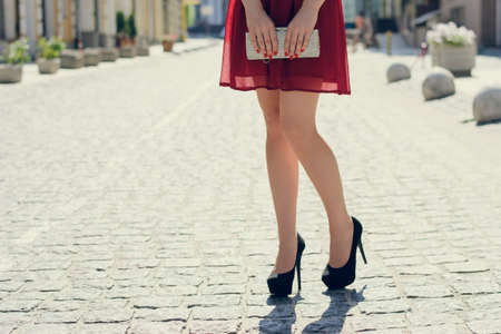 Close up photo of woman's logh legs against view of the city. She is wearing red dress and black high-heels and holding bag in handsの写真素材