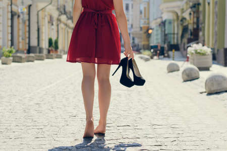 Woman in elegant red dress holding her high heel shoes in hands and walking barefoot, coming back home after party in the morning; view from back; city on the backgroundの写真素材