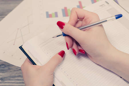 Concept of workplace. Close up of woman's hand writing information in diary with graph on backgroundの写真素材