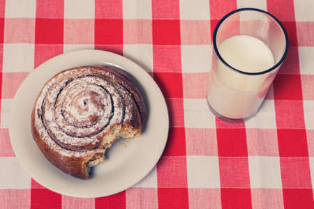 Top view photo of bitten cinnamon roll on a plate and glass of milk on checkered tableclothの写真素材