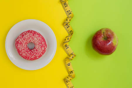 Diet or calories? Concept of unhealthy lifestyle. Tasty donut, fresh juicy apple, tape measure on yellow and green background. Top viewの写真素材