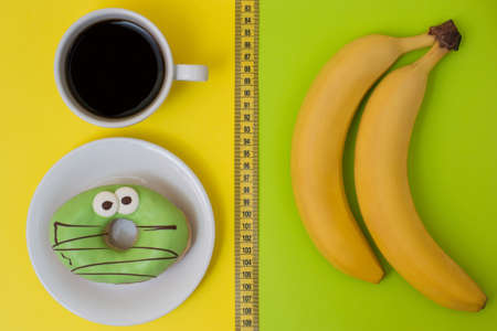 Concept of unhealthy eating and snack. Delicious donut and cup of strong coffee isolated on yellow and fresh tasty bananas isolated on green background. Top view photoの写真素材