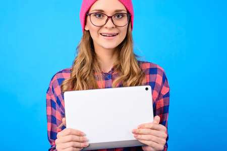 Close up portrait of excited cheerful satisfied teen girl wearing casual checkered shirt pink hat nerd eyewear hugging white with camera digital tablet isolated on blue background copy-spaceの写真素材