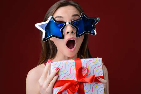 Concept of happiness when receiving presents on Christmas. Portrait of happy surprised crazy woman with open mouth. She is wearing big funny sunglasses and holding present box.の写真素材