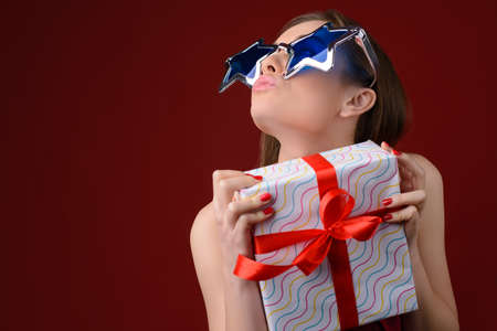 Funny happy woman in big sunglasses sending air-kiss and holding present box in her hands. Isolated on red backgroundの写真素材