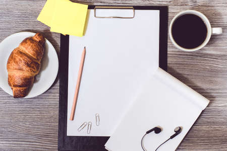 Concept of office accessorise lying on wooden desktop: clipboard, croissant, cup of coffee, earphonesの写真素材