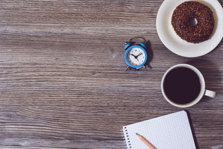Top view with copyspace. Office workplace with cup of coffee, donut, alarm clock, notepad on wooden...