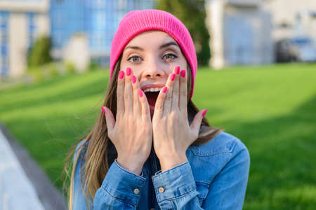 Happy joyful surprised  teenage girl in pink hat, with pink nails, dressed in jeans shirt covering her mouth with palmsの写真素材