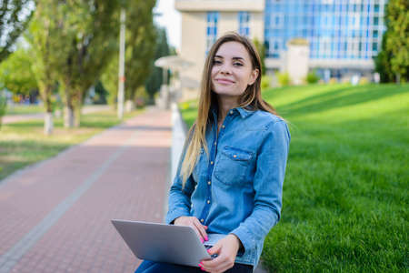 Smiling happy girl in jeans shirt using laptop for communication with friends via the Internet. Modern building is on the backgroundの写真素材
