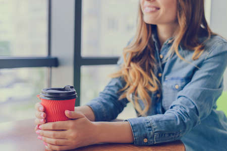 Pretty smiling woman sitting near the window in a cafe in casual clothing drinking fresh tasty coffee and having a conversation with her friendの写真素材