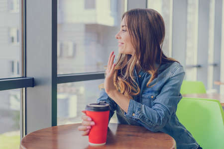 Smiling young girl in jeans shirt drinking coffee, sitting in a cafe and waving to her friendの写真素材
