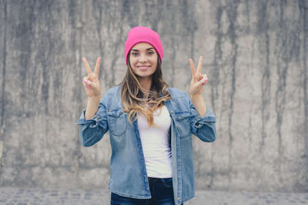 Happy joyful smiling hipster girl in casual clothes and pink hat standing against gray wall and showing victory signの写真素材