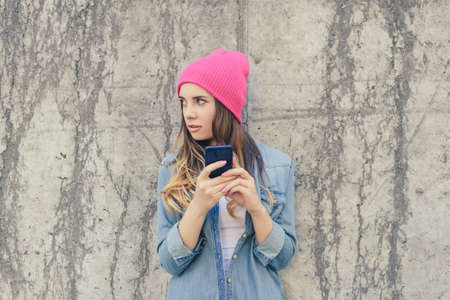 Jealous woman checking her boyfriend's mobile phone. Woman reading secret sms on her mobile phone. She is dressed in casual clothes, pink hat. Close up photo, grey stone backgroundの写真素材