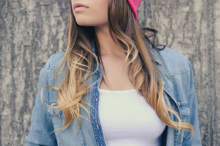 Stylish hipster woman with long hair in jeans clothing and pink hat, in white t-shirt standing against concrete wall on the street. Clopped, close-up photo, empty space on t-shirtの写真素材