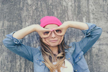 Woman having fun. Woman making binoculars using her hands. She is dressed in jeand shirt and pink hat. Grey backgroundの写真素材