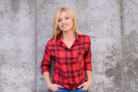 Woman smiling at camera. Woman with blonde hair, dressed in red checkered shirt and jeans standing against grey wall. Portrait, close up photoの写真素材