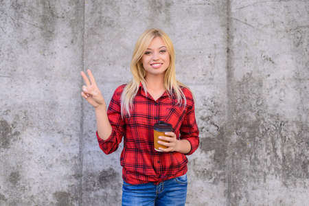 Smiling happy woman with blonde hair dressed in casual clothes is showing victory sign and holding morning coffeeの写真素材