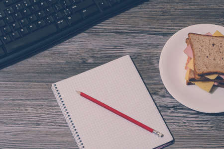 Break at work. Tasty cheese sandwich, notebook and pencil with keyboard on gray wooden background. Accent on a notebook. Vertical view, vintage effectの写真素材