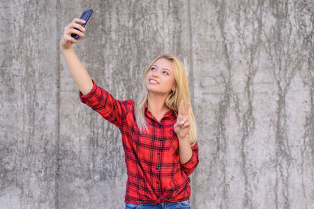 Woman in casual clothes with beaming smile taling selfie on her smartphone and showing v-signの写真素材