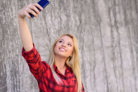 Young smiling girl with beautiful face taking self-portrait on her smartphone. She has blonde hair, beaming smile. She is wearing red checkered shirt. Close up portrait, grey wall on the backgroundの写真素材
