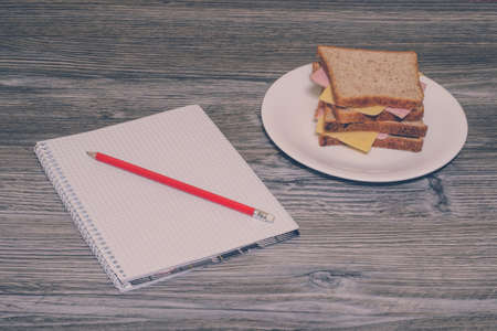 Dinner at work. Sandwich with ham and sausage on a white plate, notebook with pencile on it. Horizontal view, gray wooden background.の写真素材